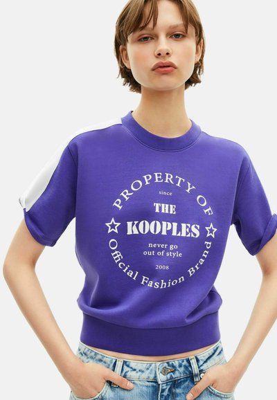 Młoda osoba ubrana w fioletowy t-shirt z krótkim rękawem z białym napisem "WŁASNOŚĆ KOOPLES" oraz niebieskie dżinsy.
