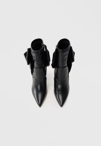 Bottines en cuir noir avec un bout pointu, dotées d'une fermeture à glissière et d'une texture froncée à l'arrière, ornées de nœuds décoratifs.