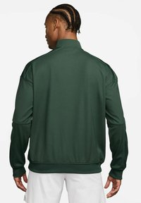 Uomo con capelli intrecciati che indossa una giacca maniche lunghe verde scuro e pantaloni bianchi, rivolto con la schiena verso la fotocamera su uno sfondo semplice.