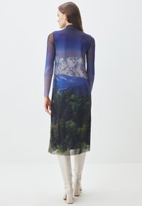 Abito trasparente a maniche lunghe con una stampa di paesaggi naturali, comprendente montagne e foreste nei toni di blu, verde e bianco. Abbinato a stivali bianchi fino al ginocchio.