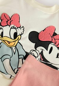 Bomullströja med tryck av tecknade figurer av en anka och en mus, prydd med rosa texturerade rosetter. Bredvid finns ett ribbat rosa tyg.