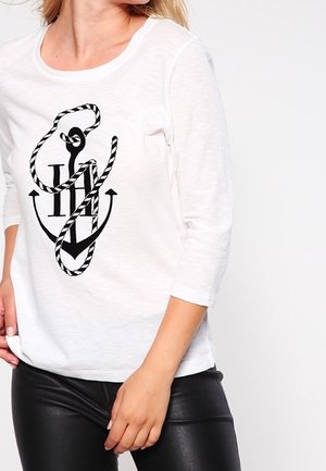 Femme portant une chemise blanche à manches longues avec un motif ancre et corde noir, associée à un pantalon en cuir noir.