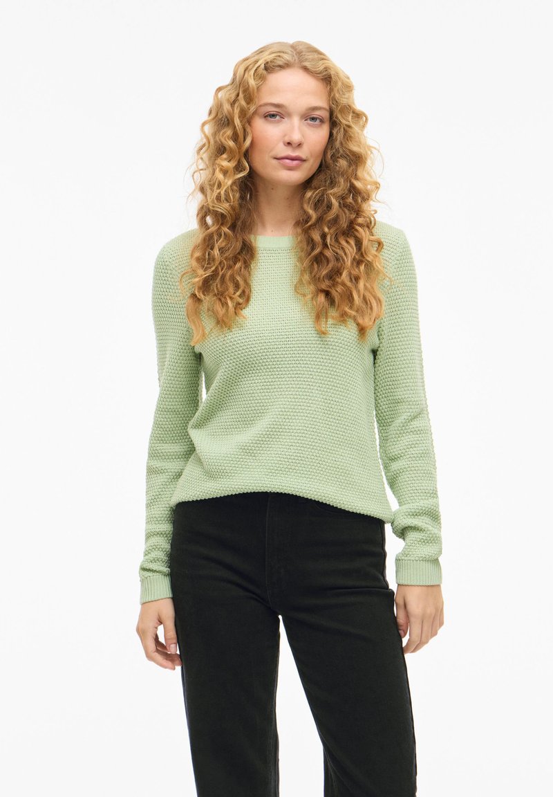 Ανοιχτό πράσινο πλεκτό πουλόβερ με υφασμένο μοτίβο και ριμπ μανσέτες, συνδυασμένο με μαύρο παντελόνι. Το πουλόβερ έχει στρογγυλή λαιμόκοψη και άνετη εφαρμογή.