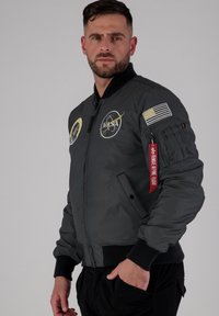 Giacca bomber grigia con polsini e orlo a coste, toppe NASA, bandiera americana sulla manica e etichetta rossa "Rimuovere prima del volo". Texture liscia.