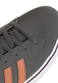 Baskets grises avec des lacets gris foncé, deux bandes orange sur le côté, et une semelle blanche avec une fine ligne bordeaux.