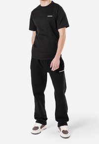 Svart bomull t-shirt och svarta sweatpants, båda med vita brandingdetaljer. Skor med en blandning av beige och bruna nyanser.