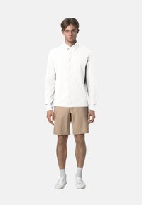 Camicia bianca a maniche lunghe con colletto, pantaloni corti beige e sneakers bianche. Tessuto liscio, design semplice e tasche frontali visibili sui pantaloni.