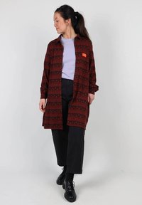 Chemise rouge à motifs avec des manches longues, design à boutons, et une étiquette orange. Portée par-dessus un haut violet clair et associée à un pantalon et des chaussures noirs.
