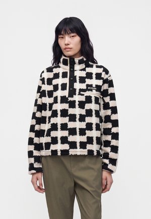 Femme portant un pull en polaire à carreaux noir et blanc avec un col boutonné et un pantalon vert olive sur un fond blanc uni.