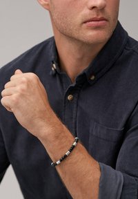 Homme portant une chemise boutonnée bleu foncé avec un bracelet en perles noir et blanc à son poignet gauche, bras levé près de la poitrine.