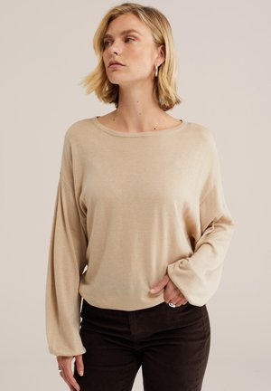 Jeune femme aux cheveux blonds courts portant un pull beige manches longues et un pantalon foncé, posant avec une main dans la poche, regardant sur le côté.