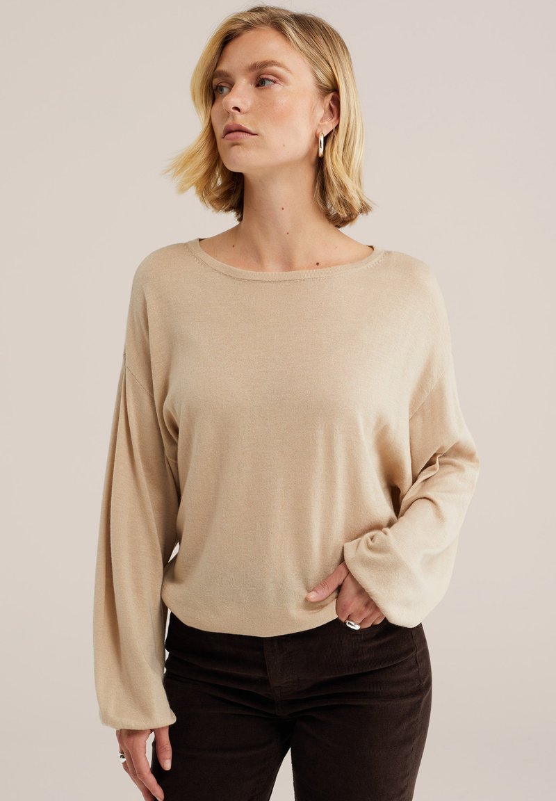 Jeune femme aux cheveux blonds courts portant un pull beige manches longues et un pantalon foncé, posant avec une main dans la poche, regardant sur le côté.