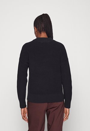 Osoba z warkoczem związanym w wysoki kucyk, ubrana w czarny dzianinowy sweter i brązowe spodnie, stojąca twarzą do jasnoszarego tła.