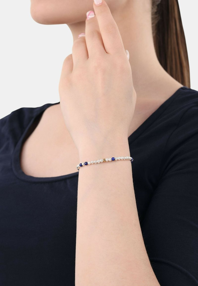 Pulsera de cuentas metálicas con cuentas alternas blancas, azules y doradas, llevada en una muñeca delgada sobre una camiseta oscura. Textura suave.