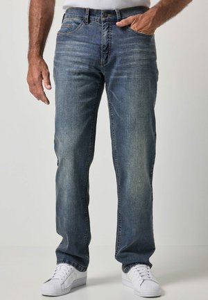 Homme portant un jean bleu délavé et des baskets blanches, debout avec une main dans la poche sur un fond uni.