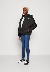 Veste doudoune noire avec capuche, présentant un design matelassé et un petit logo blanc. Portée sur un haut gris et assortie à un jean bleu et des baskets noires.