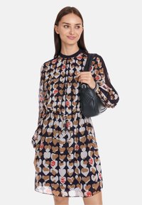 Kleid mit Blumenmuster auf navy Hintergrund, versehen mit Herzen in Braun und Weiß. Lange Ärmel, geraffte Taille, kombiniert mit einer kleinen schwarzen Handtasche.