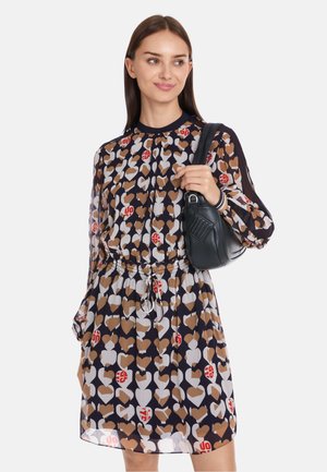 Kleid mit Blumenmuster auf navy Hintergrund, versehen mit Herzen in Braun und Weiß. Lange Ärmel, geraffte Taille, kombiniert mit einer kleinen schwarzen Handtasche.