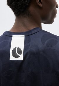 Οπίσθια όψη ενός ατόμου που φοράει μπλε σκούρο πουκάμισο με λευκή κάθετη ρίγα και μερικώς ορατό λογότυπο σε σχήμα κύκλου στο άνω μέρος της πλάτης.