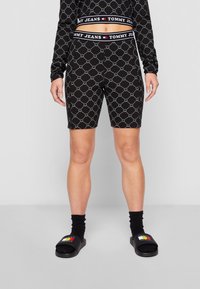 Pantaloni ciclistici neri con un motivo geometrico e vita elasticizzata, abbinati a sandali neri e calzini alla caviglia, il tutto su uno sfondo neutro.