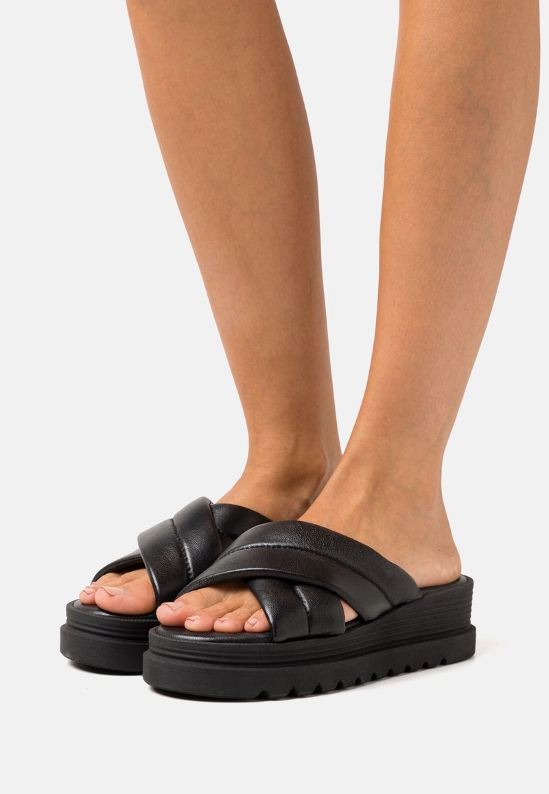 Pieds portant des sandales plates-formes noires avec des brides en cuir épaisses croisées et des semelles texturées sur un fond clair.