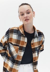 Camisa de franela a cuadros en tonos marrones, blancos y negros. Presenta un estilo de botón, cuello y un bolsillo en el pecho. Lleva una camiseta negra debajo.