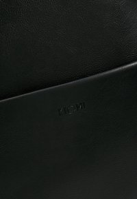 Zwarte leren item met het reliëf 'KIOMI' logo; heeft een gladde textuur en een minimalistisch ontwerp, dat de rijkdom van het materiaal benadrukt.