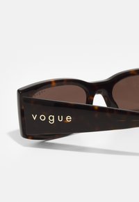 Čūskastīšanas saulesbrilles ar brūnajiem stikliem, ar drosmīgu, leņķisku dizainu un zelta "vogue" logo uz tempļa. Gluda tekstūra.