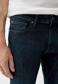 Donkerblauwe spijkerbroek met een slim fit, voorzien van een voorzak met metalen klinknagels en zichtbare stiksels. Close-up van het taillegebied.