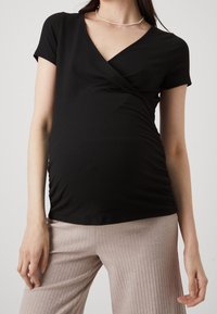 Černý žebrovaný těhotenský top s výstřihem do V a krátkými rukávy, který má zdobení na bocích pro tvarování a pohodlí. V kombinaci se světle žebrovanými kalhotami.
