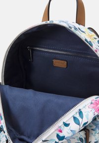 Sac à dos à motif floral avec un extérieur blanc, des bretelles en cuir marron, et un intérieur bleu marine comprenant une poche zippée et une doublure texturée.