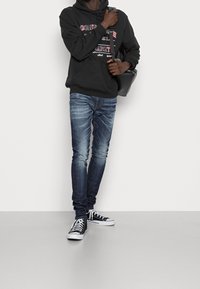 Personne portant un sweat à capuche noir avec du texte, un jean bleu délavé, et des baskets noires montantes, tenant la sangle d'un sac à bandoulière noir devant un fond blanc.