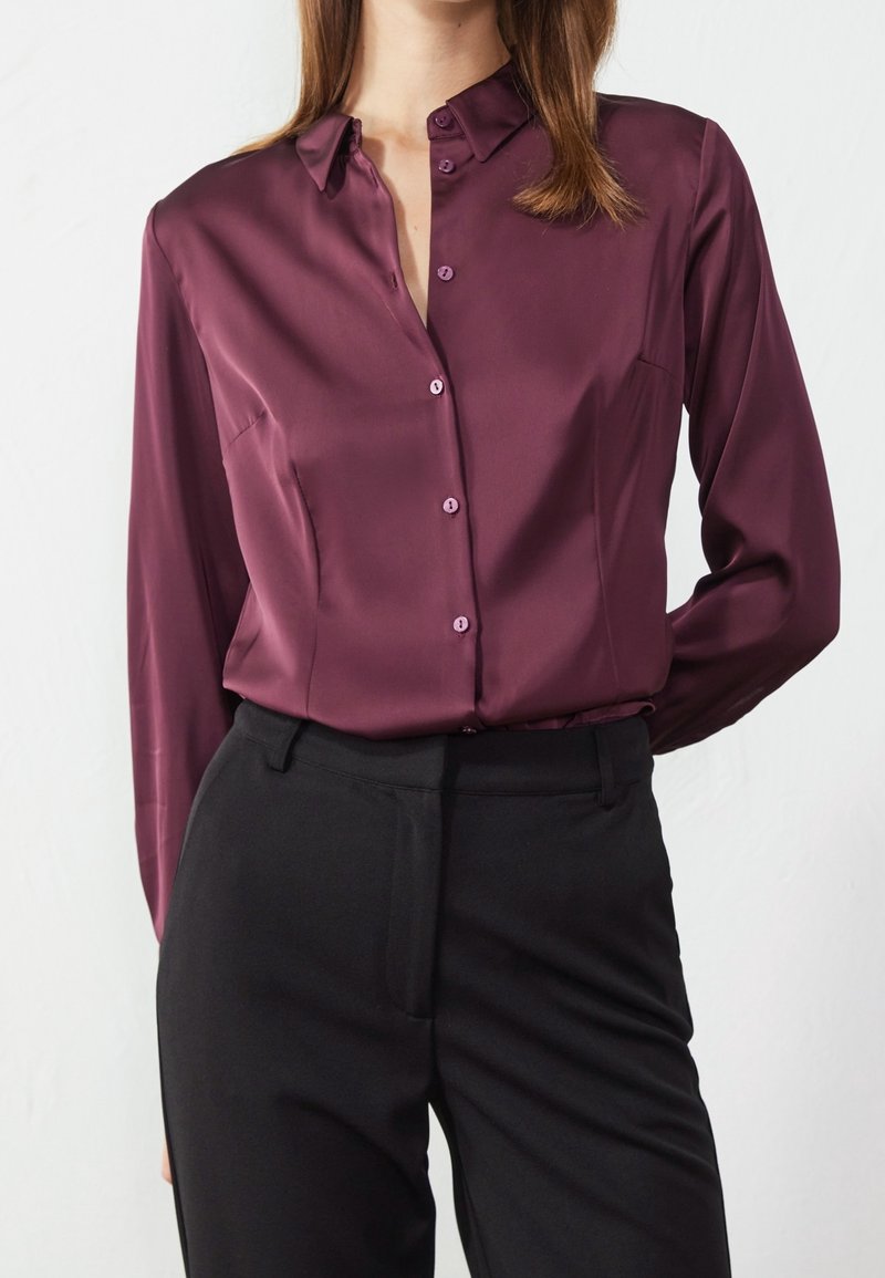Femme portant un chemisier en satin violet à manches longues, boutonné et rentré dans un pantalon noir taille haute, sur un fond blanc uni.