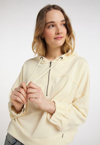 Kremowa bluza z kapturem z zamkiem błyskawicznym z przodu, elastycznymi mankietami i sznurkiem do regulacji. Cecha to luźny krój i miękki, teksturowany materiał.