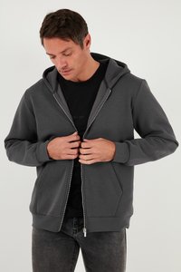 Grå zip-up hoodie med frontdragkedja, två sidofickor och fodrad huva. Tyget verkar slätt med en mjuk textur. Bärs över en svart tröja.