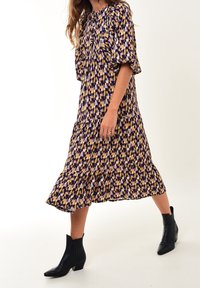 Vestido fluido de longitud media en estampado geométrico de púrpura, amarillo y lila. Mangas cortas abullonadas, dobladillo en capas, combinado con botas de tobillo negras.
