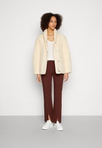 Casaco puffer creme com gola alta, combinado com calças largas castanhas com fendas e ténis brancos. Texturas suaves e linhas limpas.