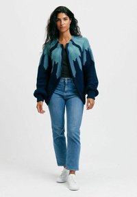 Stickad cardigan med blågrön bergsmönster, öppen framdel, breda ärmar och avslappnad passform. Matchas med ljusa denimbyxor och vita sneakers.