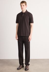 Mladý muž stojí, oblečený v tmavohnedom polo tričku s krátkym rukávom, zodpovedajúcich nohaviciach a hnedých kožených dress shoes, na neutrálnom pozadí.
