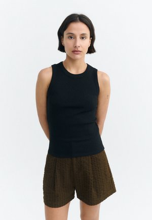 Young woman with short dark hair wearing a sleeveless black top and brown textured shorts, standing with hands behind back on white background.