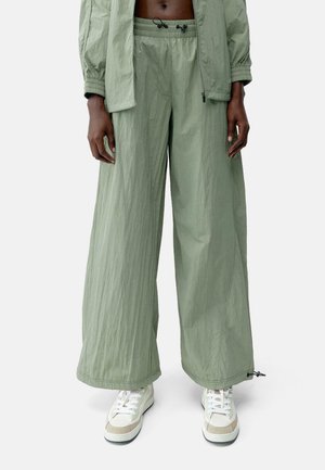 Personne portant un pantalon vert clair à jambes larges avec une taille élastique et des baskets blanches sur un fond uni.