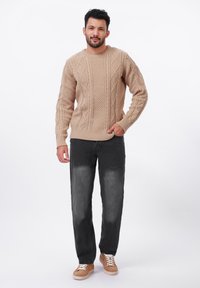 Pull en maille beige avec un motif en torsades, associé à un pantalon tapered gris foncé et des baskets beige avec des lacets blancs. Tenue simple et décontractée.
