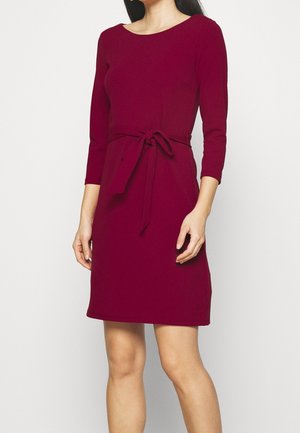 Femme portant une robe bordeaux à manches longues, longueur genou, avec une ceinture nouée à la taille, debout devant un fond clair uni.