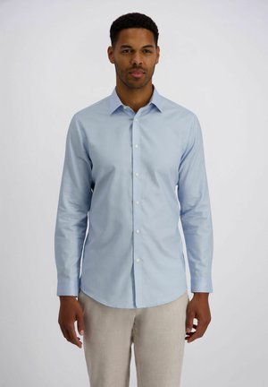 Man draagt een nauwsluitend lichtblauw overhemd met lange mouwen en knopen, en beige broek, staand tegen een effen achtergrond.