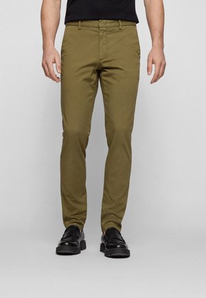 Mann trägt slim-fit olivgrüne Hose und schwarze Loafers, steht vor einem einfachen hellgrauen Hintergrund.