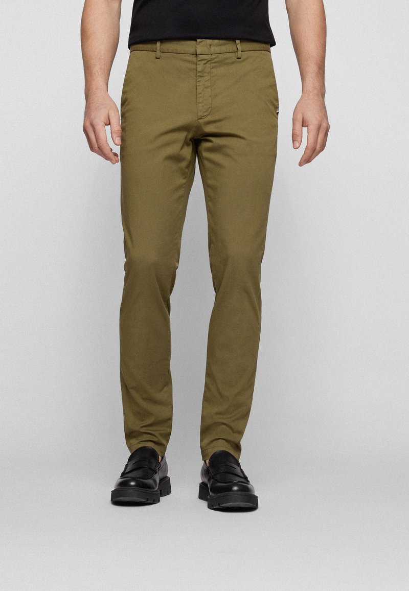 Homme portant un pantalon slim vert olive et des mocassins noirs, debout devant un fond gris clair uni.