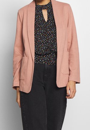 Kobieta w jasnoróżowej marynarce na czarnej bluzce w kolorowe kropki i czarnych spodniach z wysokim stanem.