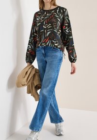 Personne portant un jean bleu, un haut à manches longues à motifs, des baskets argentées, tenant une veste beige devant un fond uni blanc et beige.