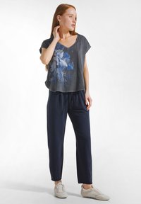 Młoda kobieta z rudymi włosami ubrana w ciemnoszarą koszulkę z kwiatowym nadrukiem, granatowe spodnie z sznurkiem i jasnobeżowe trampki, stojąca na białym tle.