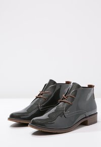 Bottines en cuir verni gris avec bouts ronds, lacets marron, semelles beige clair et détails de couture minimalistes sur les côtés.